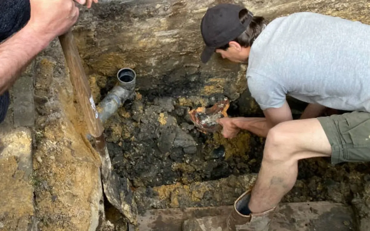 Technicians excavating and repairing an exterior drain line beside a foundation